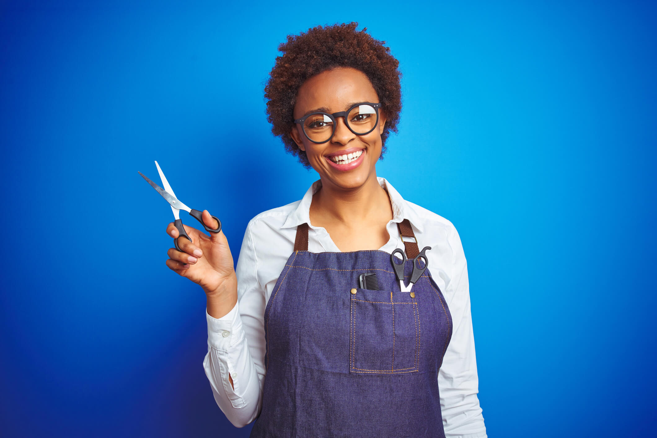 african american hairdresser woman holding scissors-over blue isolated background happy face standing smiling