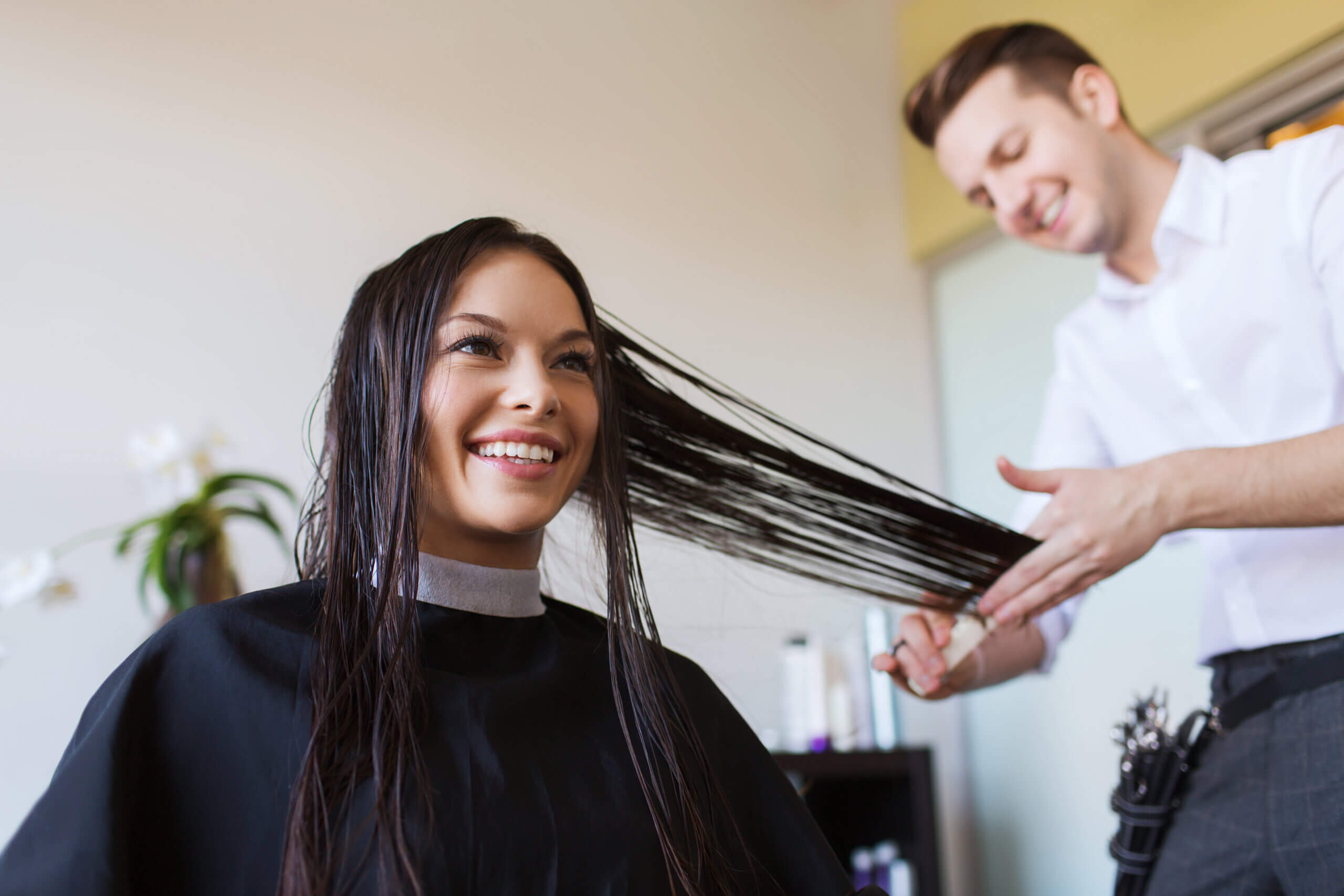 Happy woman with stylist cutting hair at salon. Beauty, hairstyle and people concept - happy young women and hairdresser cutting hair tips at salon