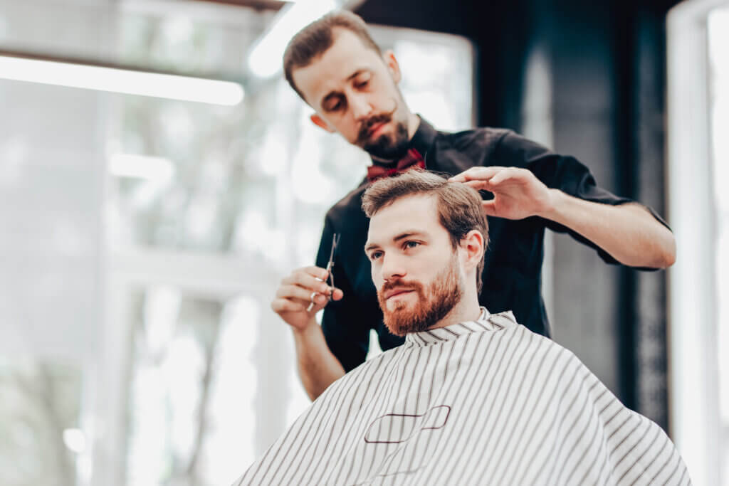 Stylish mustachioed barber dressed in a black shirt with a red bow tie scissors the hair of a young men in a barbershop.