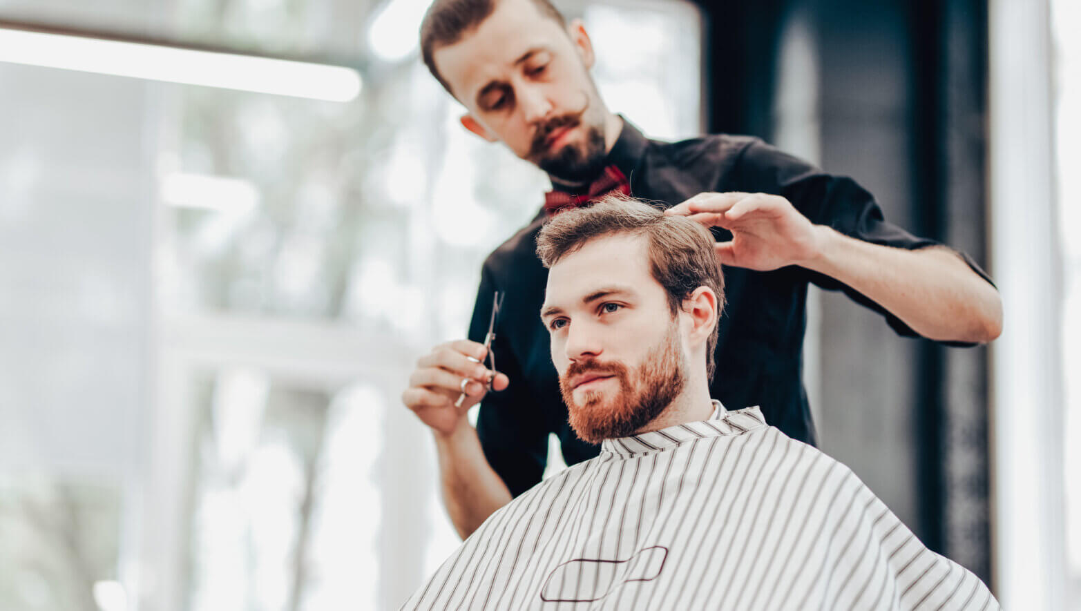 Stylish mustachioed barber dressed in a black shirt with a red bow tie scissors the hair of a young men in a barbershop.