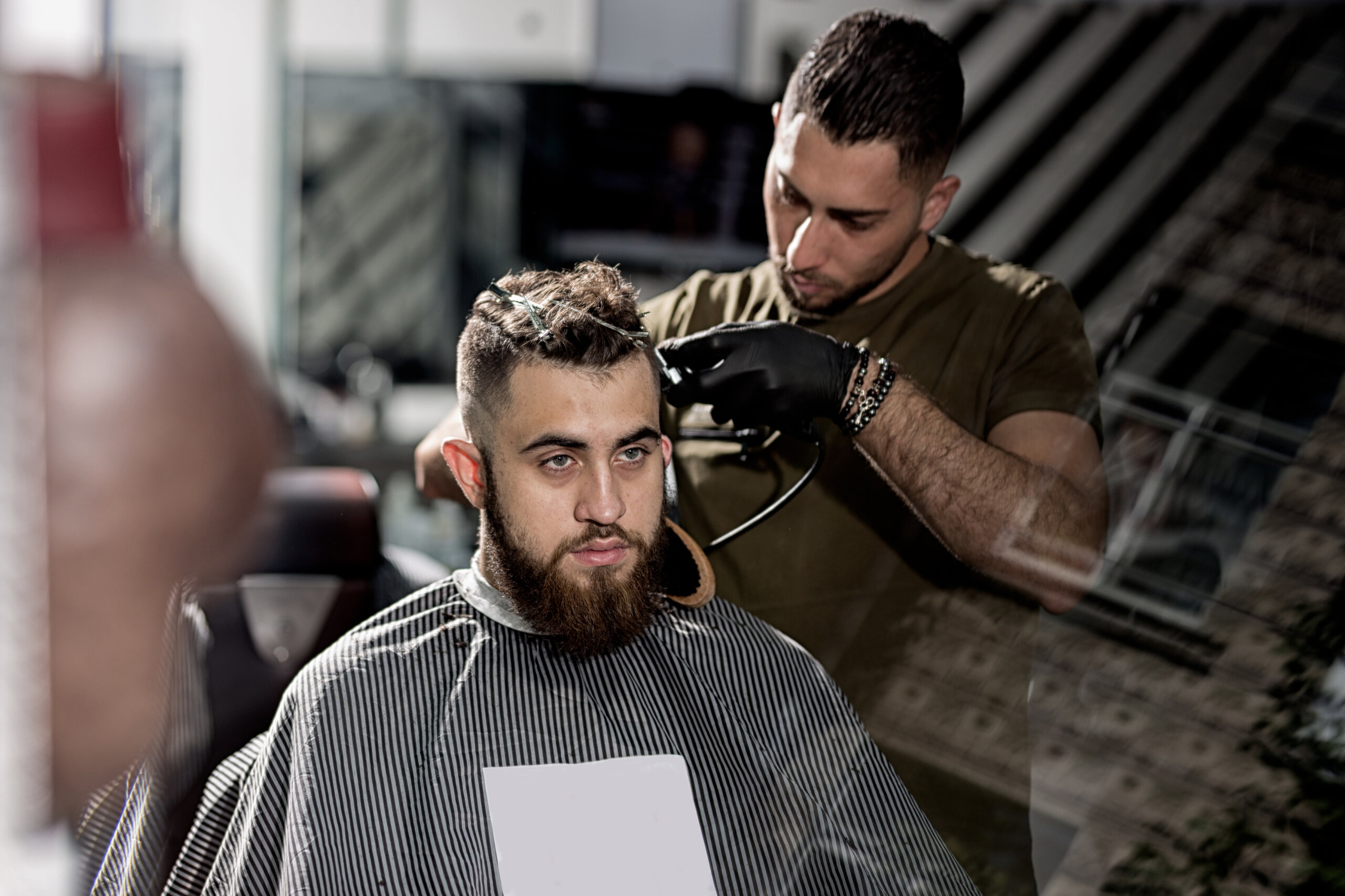 Brutal young men with beard sits at a barber shop. Barber in black gloves shaves hairs at the side.